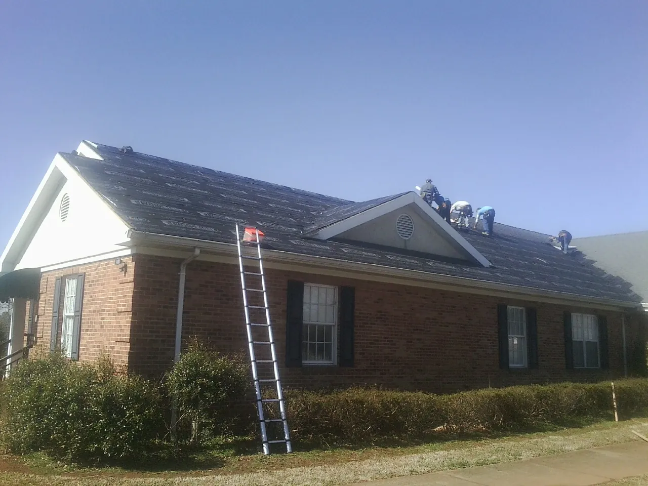 Crew tearing shingles off a brick ranch roof with a ladder set — Metal Roof Repair work by our crew in Clawson