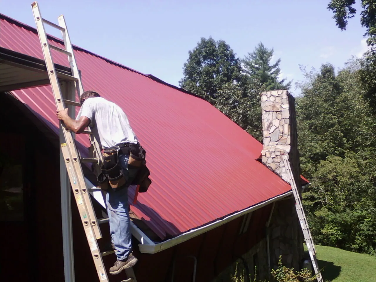 Roofer on a ladder beside a red metal roof and stone chimney — Soffit & Fascia Repair by Roofing Brothers in Clawson